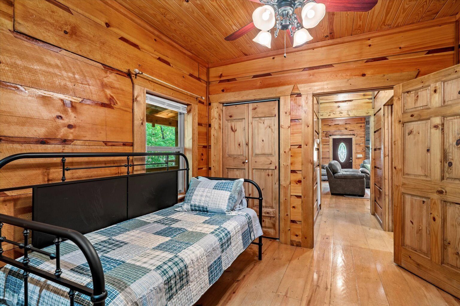 A wooden cabin interior with a bed, a ceiling fan, and a view through a window.
