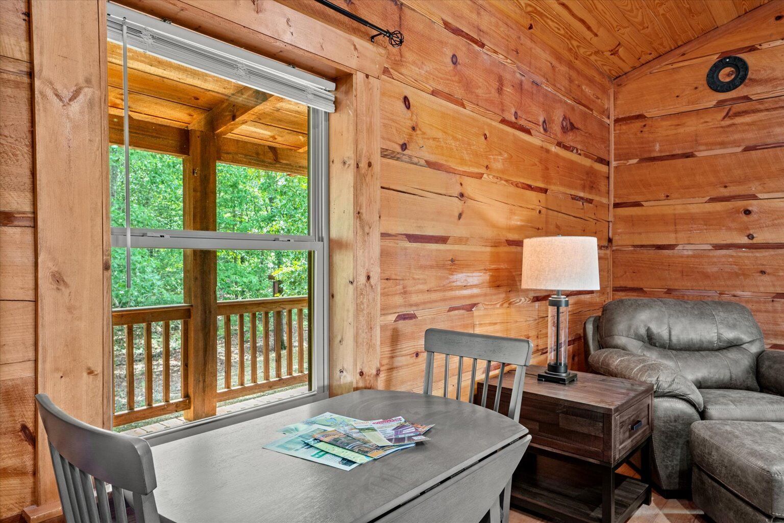 A wooden cabin interior with a dining table, chairs, a sofa, a lamp, and a window overlooking greenery.