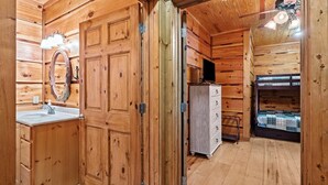 A wooden cabin interior with a wooden door, a white dresser, a bunk bed, and a ceiling fan.