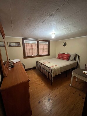 Ein Schlafzimmer mit Bett, Kommode, Nachttisch, einem Fenster mit Vorhängen und einer Deckenleuchte.
