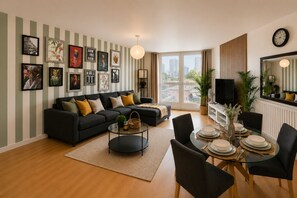 A modern living room with a sofa, coffee table, dining table, and chairs. There are framed pictures on the wall and a television on a stand. - The Galleria Suite With Free Parking (Birmingham)