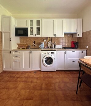 A compact kitchen with white cabinets, a microwave, a washing machine, and a dining area with a table and chairs. - The Magic Garden – Family Apartment with a Garden and Stunning Views (Teixoso)