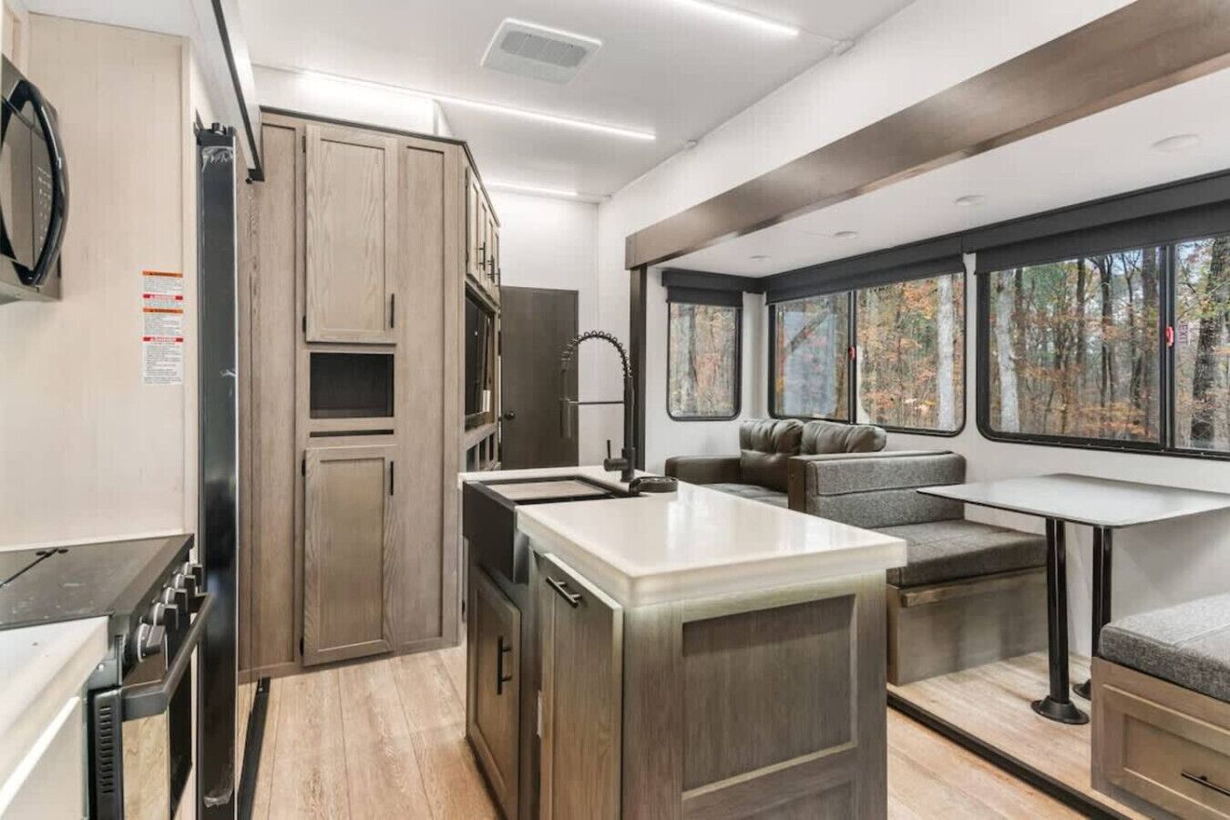 Une cuisine moderne dotée d’appareils électroménagers intégrés, un coin repas avec un banc, et de grandes fenêtres offrant une vue sur les arbres.