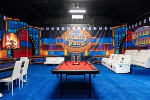 A game show set with a pool table, white sofas, and a large screen displaying 'FAMILY FEUD' and 'MOVIE NIGHT' signs. - 6 bdrm/11 beds/Indoor Pool/Hot Tub/ATVs/RV/30 acre (Ellenwood)