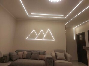 Ein modernes Wohnzimmer mit einer Couch und Sesseln, einer geometrischen Wandleuchte und einer Tür rechts.