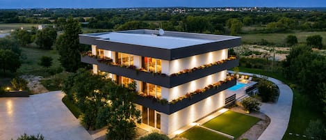 A modern multi-story house with a pool, surrounded by a landscaped garden and a view of distant mountains.