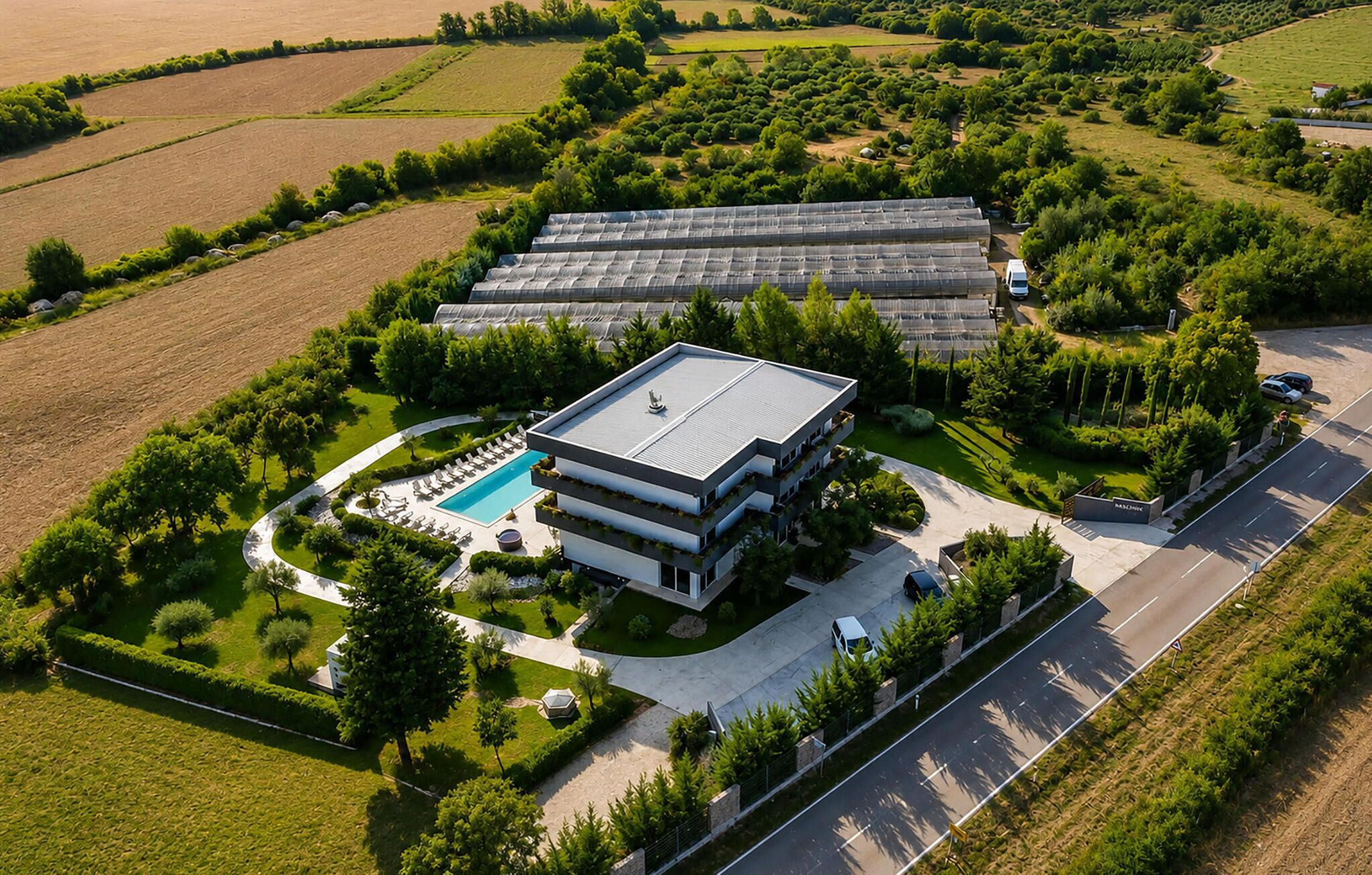 A modern building with a swimming pool, surrounded by greenery and fields.