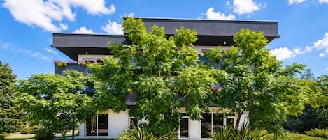 Un edificio moderno de dos pisos con techo plano, grandes ventanales y un jardín delantero ajardinado con diversas plantas y árboles.