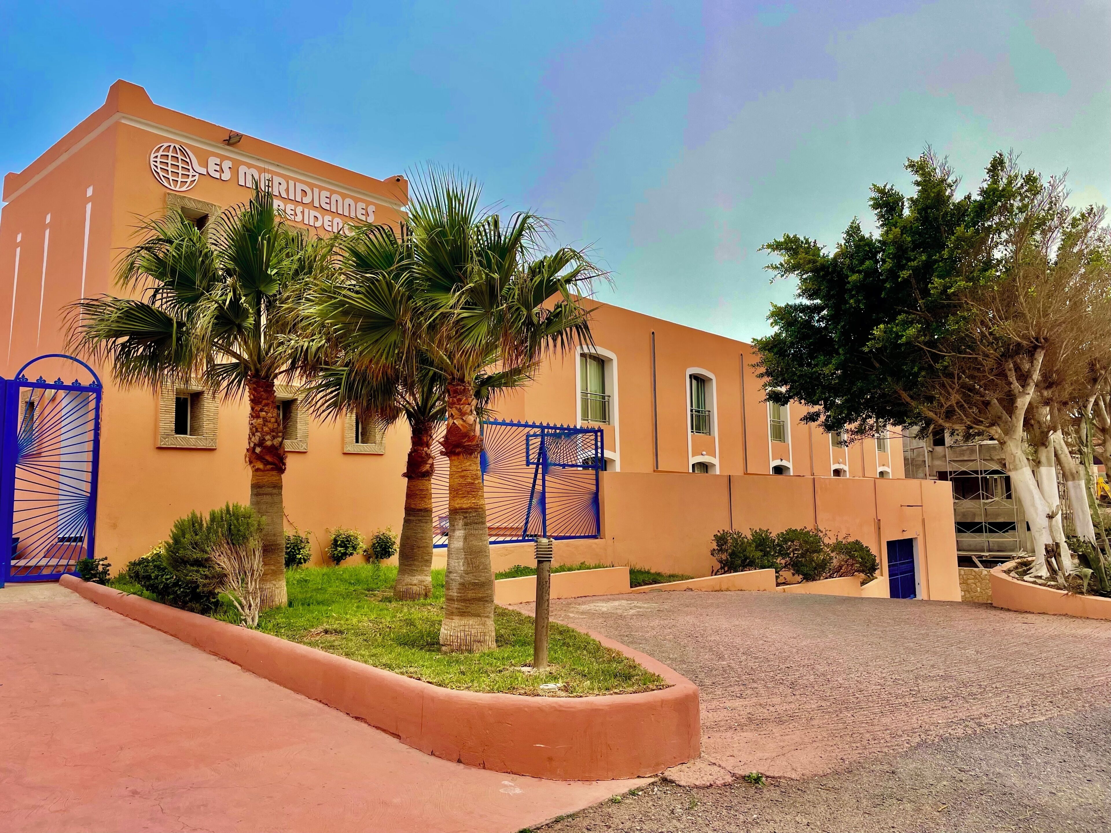 A building with a sign reading 'LES MURIDINES RESIDENCES,' palm trees, a blue gate, and a paved entrance.