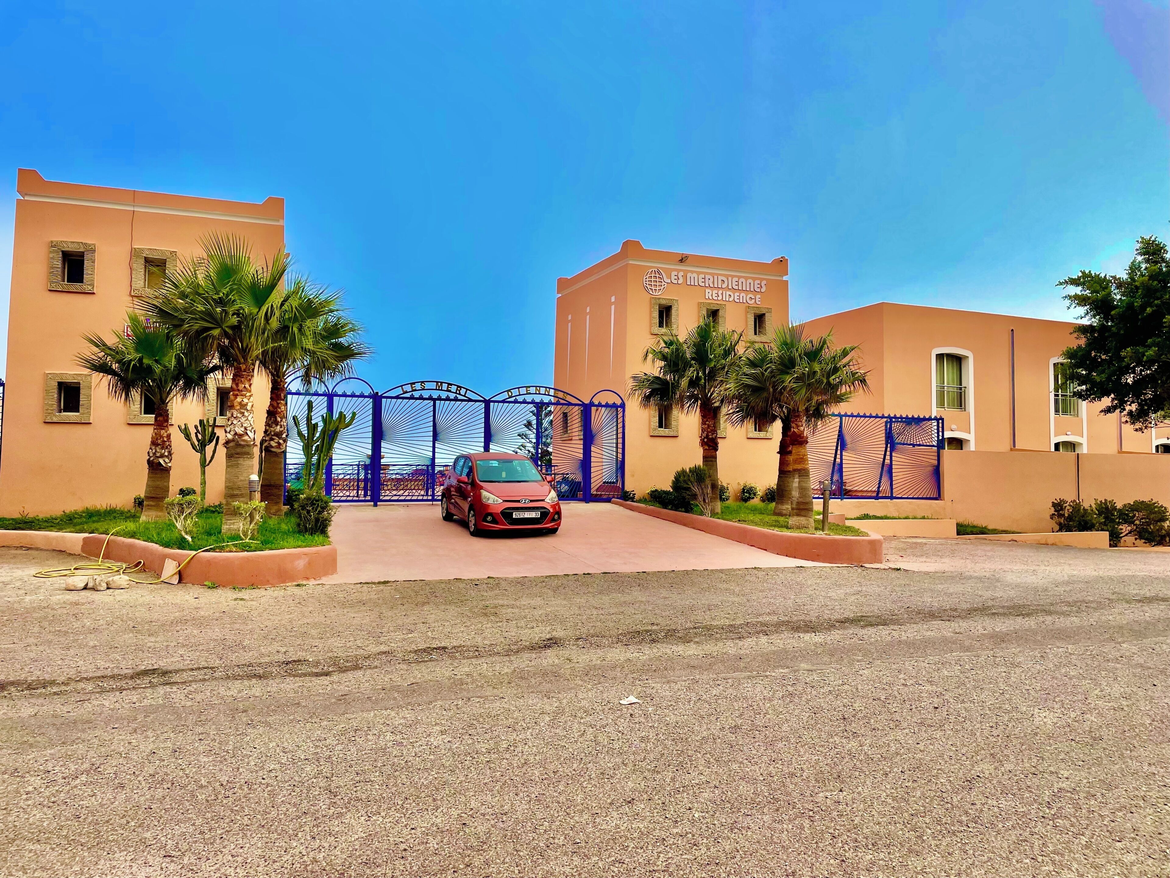 A gated entrance to a building with palm trees and a red car parked in front.