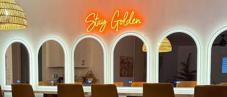 A room with a neon sign, mirrored wall, wicker chairs, and a table.