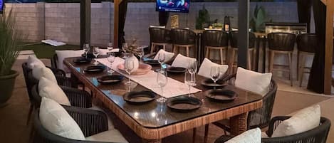 An outdoor dining area with a large wooden table, wicker chairs, and a television screen.