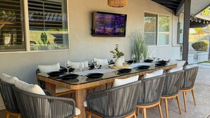 An outdoor dining area with a long table, six chairs, and a television mounted on the wall. - Pool, Putts & Paradise | Modern Lux Golden Getaway (Scottsdale)