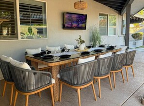 An outdoor dining area with a long table, six chairs, and a television mounted on the wall. - Pool, Putts & Paradise | Modern Lux Golden Getaway (Scottsdale)