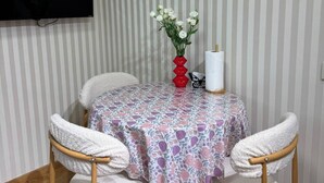 Een kamer met gestreept behang, een flatscreen televisie aan de muur, een ronde tafel met een tafelkleed met bloemenprint, twee witte stoelen, een rode vaas met witte bloemen en een witte zakdoekendoos.