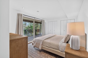 Un dormitorio con cama, mesita de noche, lámpara, armario y balcón con vista a árboles.