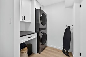 A compact laundry area with a washing machine, dryer, and ironing board. - Lakefront Luxury on Okanagan Lake | Private Beach, Dock, Hot Tub & Sauna (Kelowna)