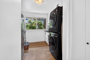 A compact laundry room with a washing machine, a dryer, and a window with a view of greenery. - Lakefront Luxury on Okanagan Lake | Private Beach, Dock, Hot Tub & Sauna (Kelowna)