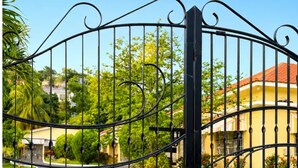 A gated entrance with a curved design, leading to a pathway surrounded by greenery and a building with a red roof. - Cozy 3-bedroom house in peaceful Kingston with AC comfort (Kingston)