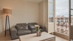 Ein modernes Wohnzimmer mit einem Ledersofa, einer Stehlampe, einem Tisch mit Blumenvase und einem großen Fenster mit Blick auf Gebäude.
