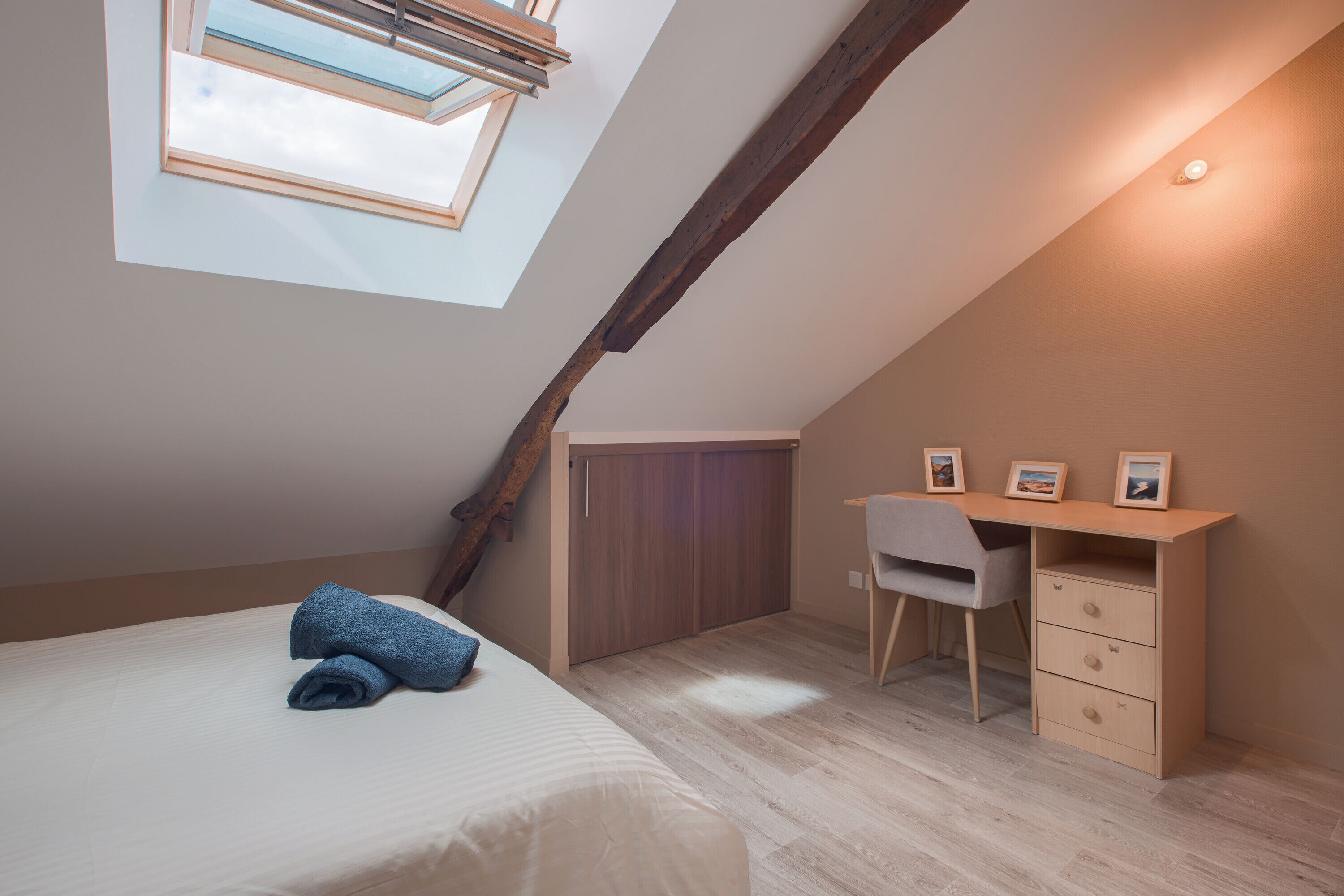 A bedroom with a sloped ceiling, a skylight, a bed with a rolled-up blanket, a desk with a chair, and a wardrobe.