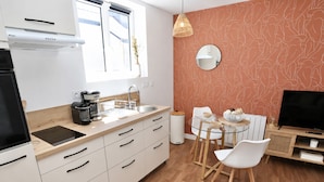 Una cocina moderna con estufa integrada, cafetera y lavamanos. Hay una zona de comedor con una mesa de vidrio y sillas blancas. Un televisor está sobre un soporte de madera y hay un espejo en la pared.