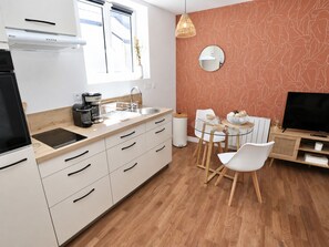 Une cuisine moderne équipée d’une cuisinière intégrée, d’une machine à café et d’un évier. On y trouve un coin repas avec une table en verre et des chaises blanches. Un téléviseur est posé sur un meuble en bois, et un miroir est accroché au mur.