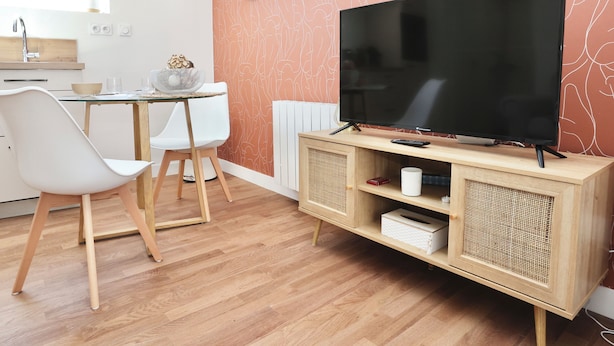 Una sala de estar moderna con un televisor de pantalla plana sobre un soporte de madera, sillas blancas, una mesa de comedor de vidrio y una pared con un patrón abstracto único.