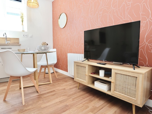 Un salon moderne avec un téléviseur à écran plat posé sur un meuble en bois, des chaises blanches, une table à manger en verre et un mur orné d’un motif abstrait original.