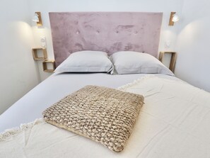 Un lit bien fait, agrémenté d’un plaid tissé et de deux oreillers, une tête de lit en bois avec des lampes fixées au mur, et un petit tabouret en bois placé à côté du lit.