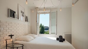 Et soveværelse med en stor seng, et glasbord med en lampe, en radiator og udsigt til grøn natur udenfor.
