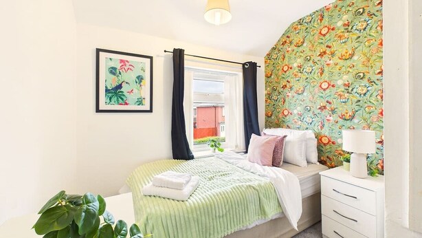 Ein Schlafzimmer mit Bett, Nachttisch, Lampe, einem Fenster mit Vorhängen, einem gerahmten Bild an der Wand und einer Pflanze.