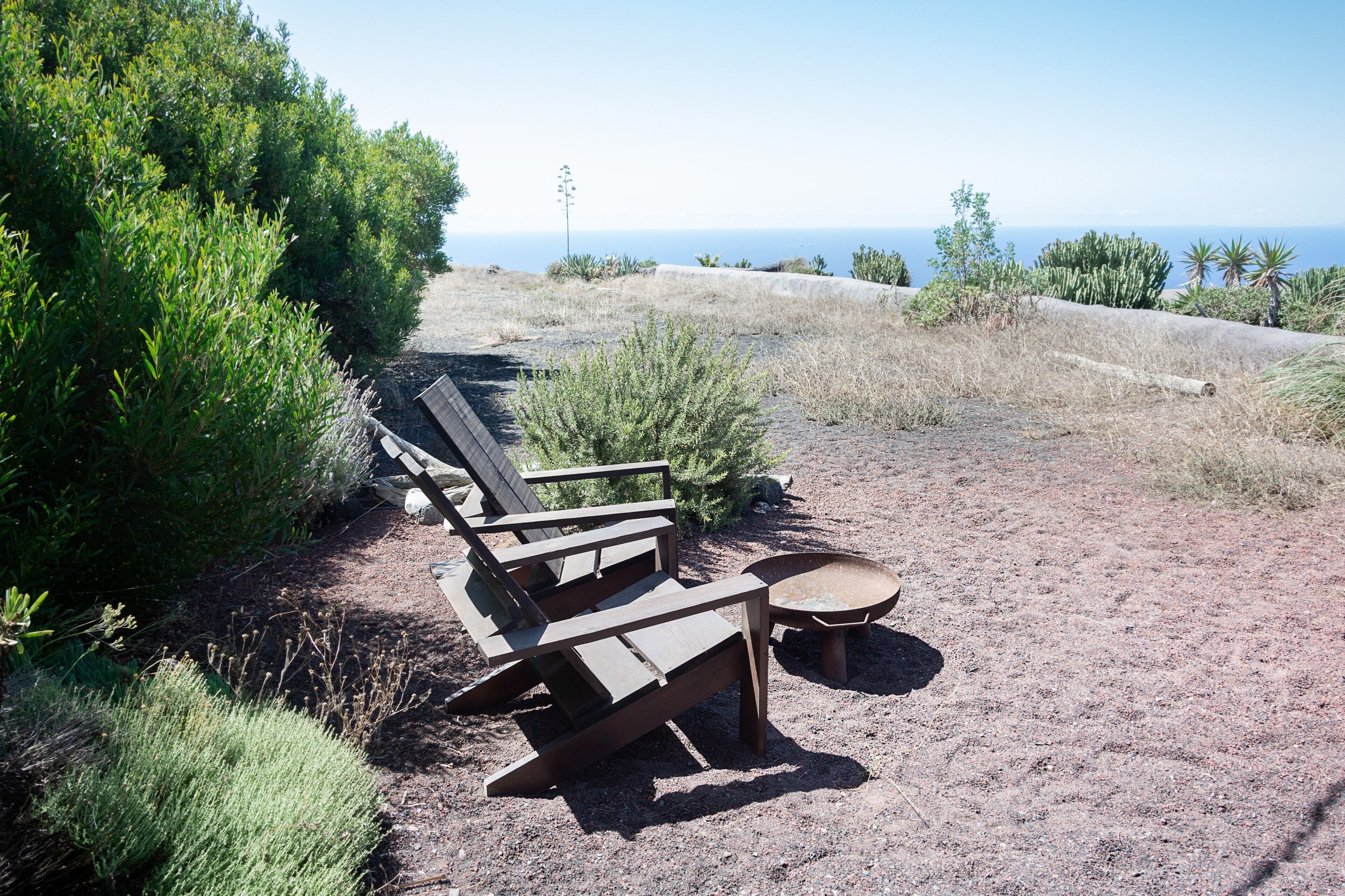 A row of wooden park benches and a wheelbarrow on a dirt path with green shrubs on one side and a view of the ocean on the other.