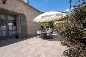 A stone building with a door and window, a table with chairs, and a white umbrella. - La Roque Vacation Rental - CANOURGUE (LA) (CANOURGUE (LA))