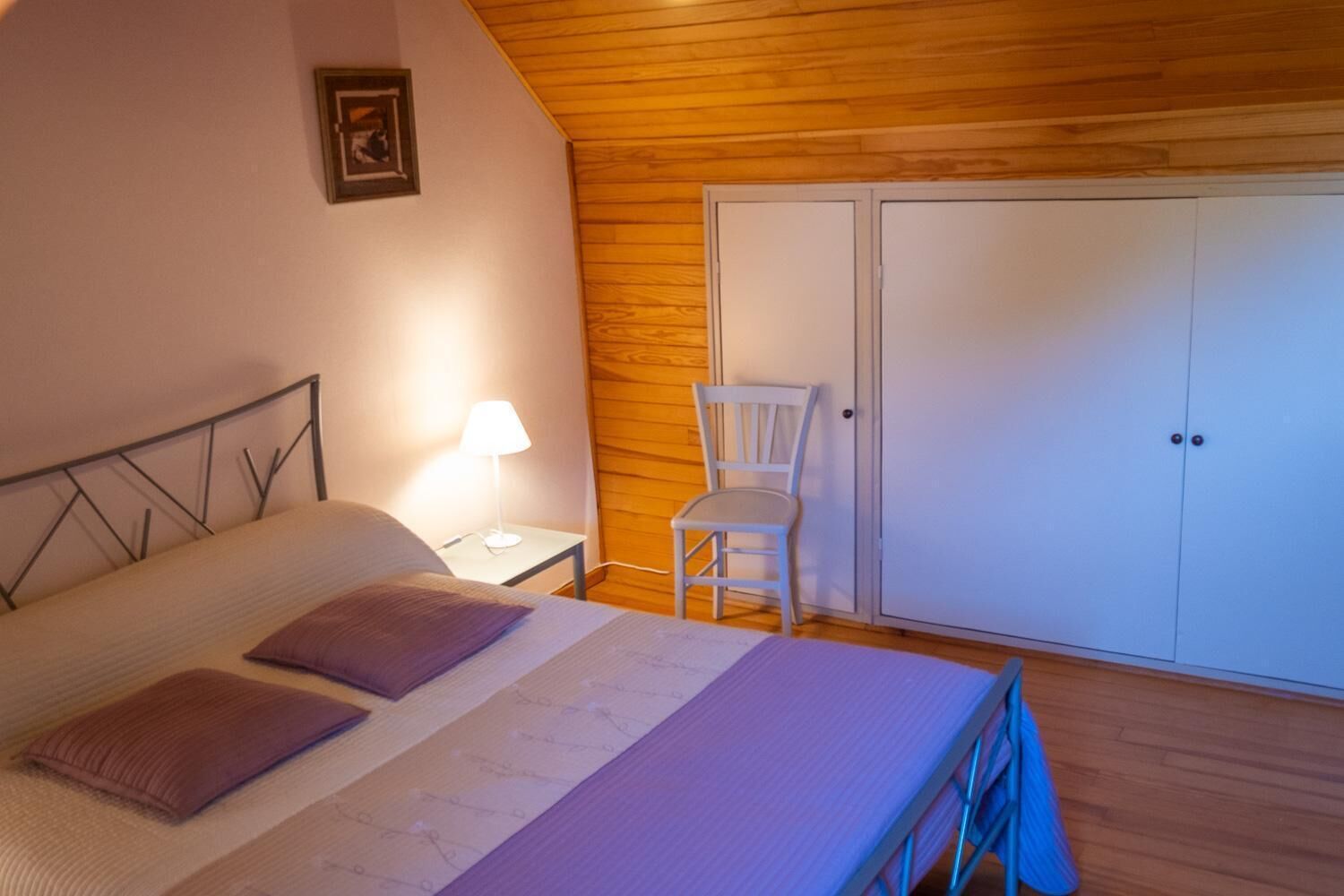 A bedroom with a bed, a nightstand with a lamp, a chair, and a wardrobe.