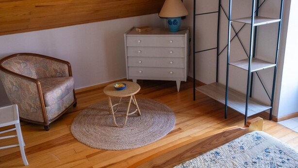 A room with a wooden ceiling, a patterned armchair, a small round table with a bowl, a white dresser, a lamp, and a metal shelving unit. - La Roque Vacation Rental - CANOURGUE (LA) (CANOURGUE (LA))