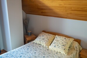 Ein Schlafzimmer mit einem hölzernen Kopfende, einem Bett mit floralem Bettzeug, einem Nachttisch mit einer Spule Faden und einer Vase mit getrockneten Ästen.