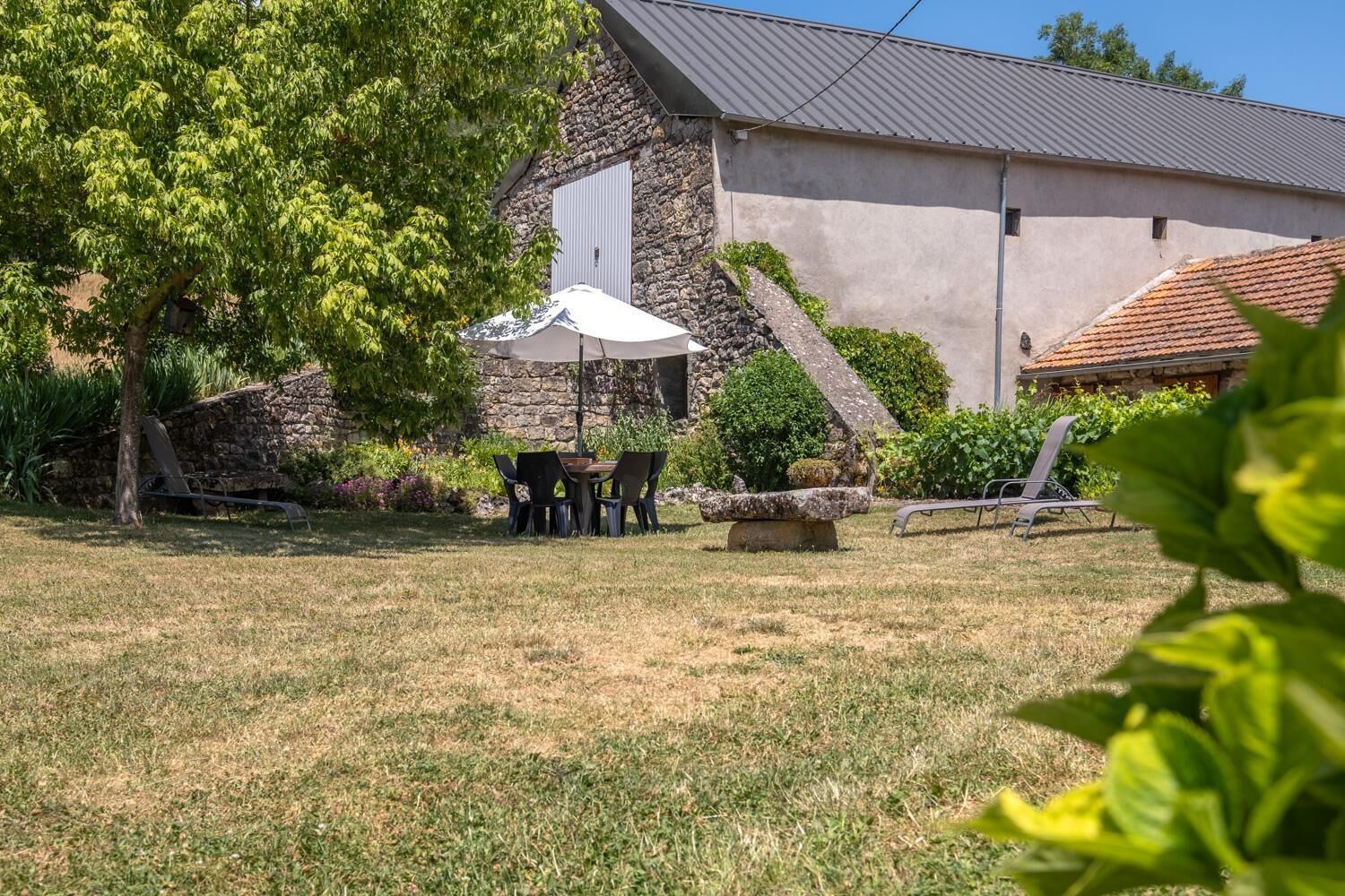 Un bâtiment en pierre doté d’un toit en pente, une terrasse avec une table et des chaises, et un jardin avec différentes espèces de plantes.