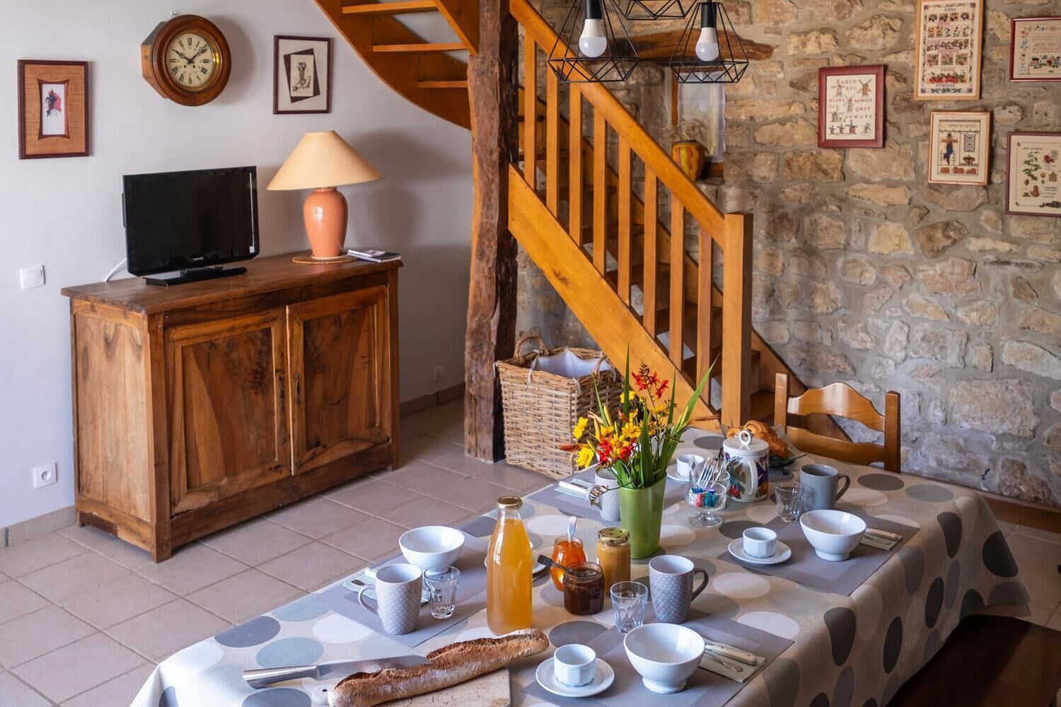 Un coin repas avec une table en bois dressée pour le petit-déjeuner, un escalier et des murs en pierre.