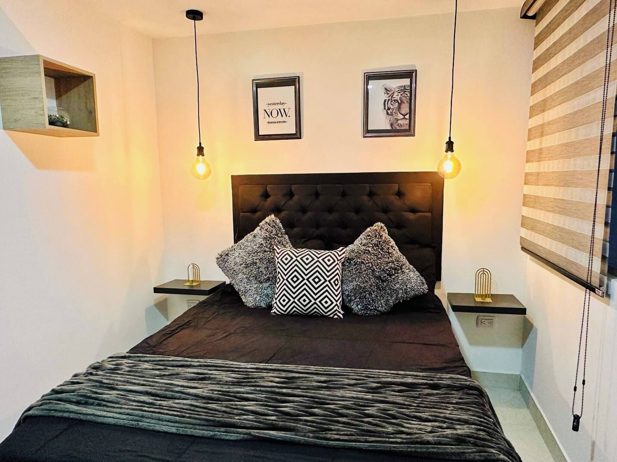 Un dormitorio con cabecera capitonada, dos cuadros enmarcados, una manta estampada y una mesita de noche con lámpara.