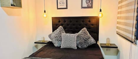 A bedroom with a tufted headboard, two framed pictures, a patterned throw blanket, and a bedside table with a lamp.