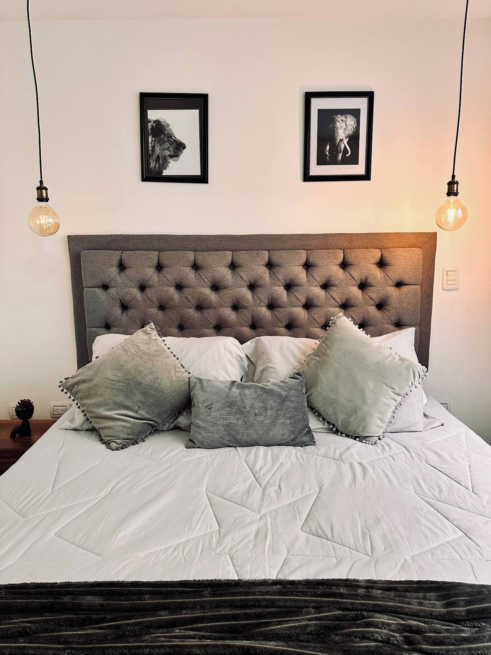 Uma cama com cabeceira estofada, várias almofadas e duas fotos emolduradas na parede.