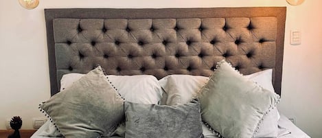 Uma cama com cabeceira estofada, várias almofadas e duas fotos emolduradas na parede.