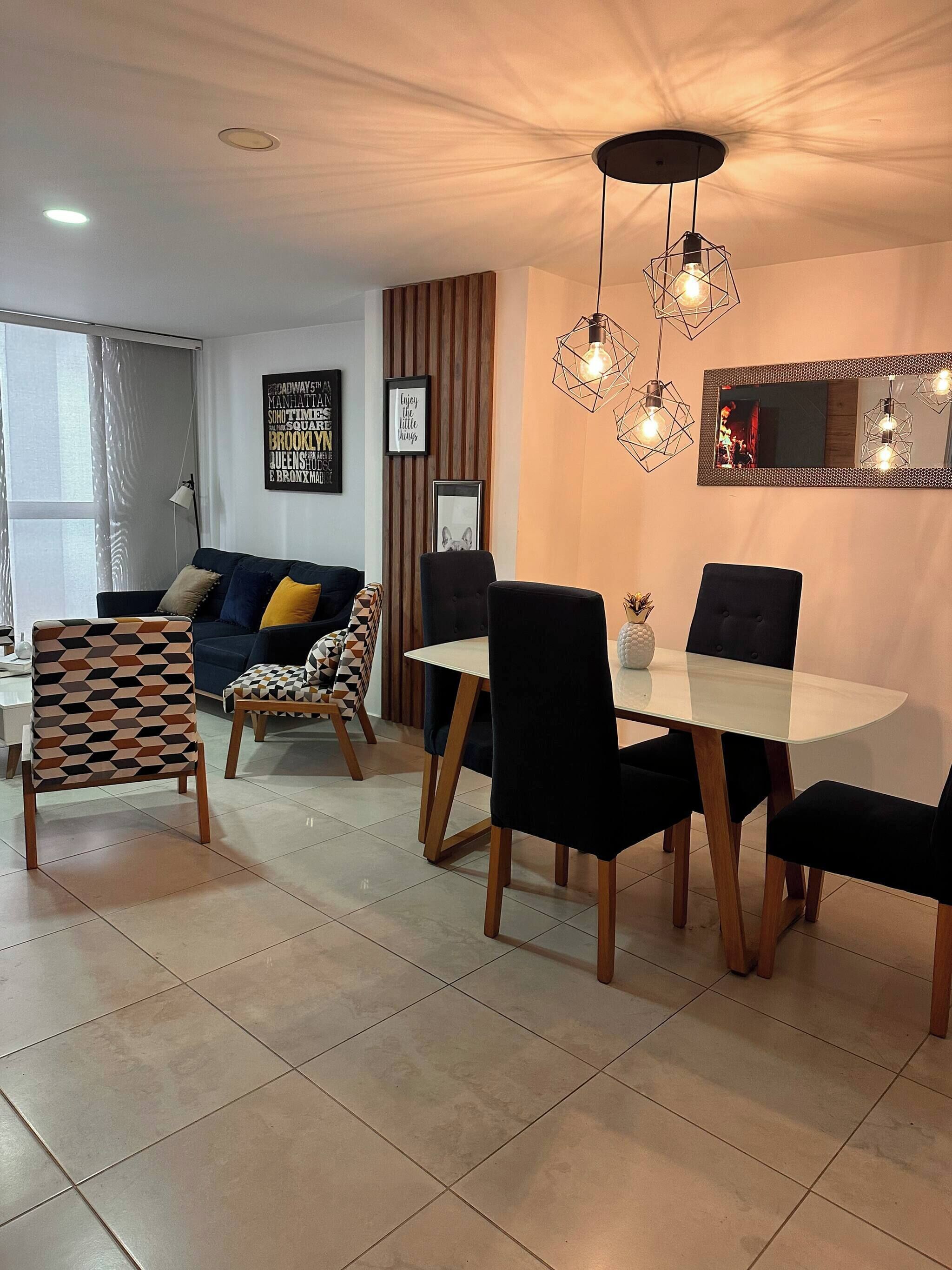 A modern living room with a sofa, a dining table with chairs, a patterned armchair, a mirror, and a ceiling light fixture.