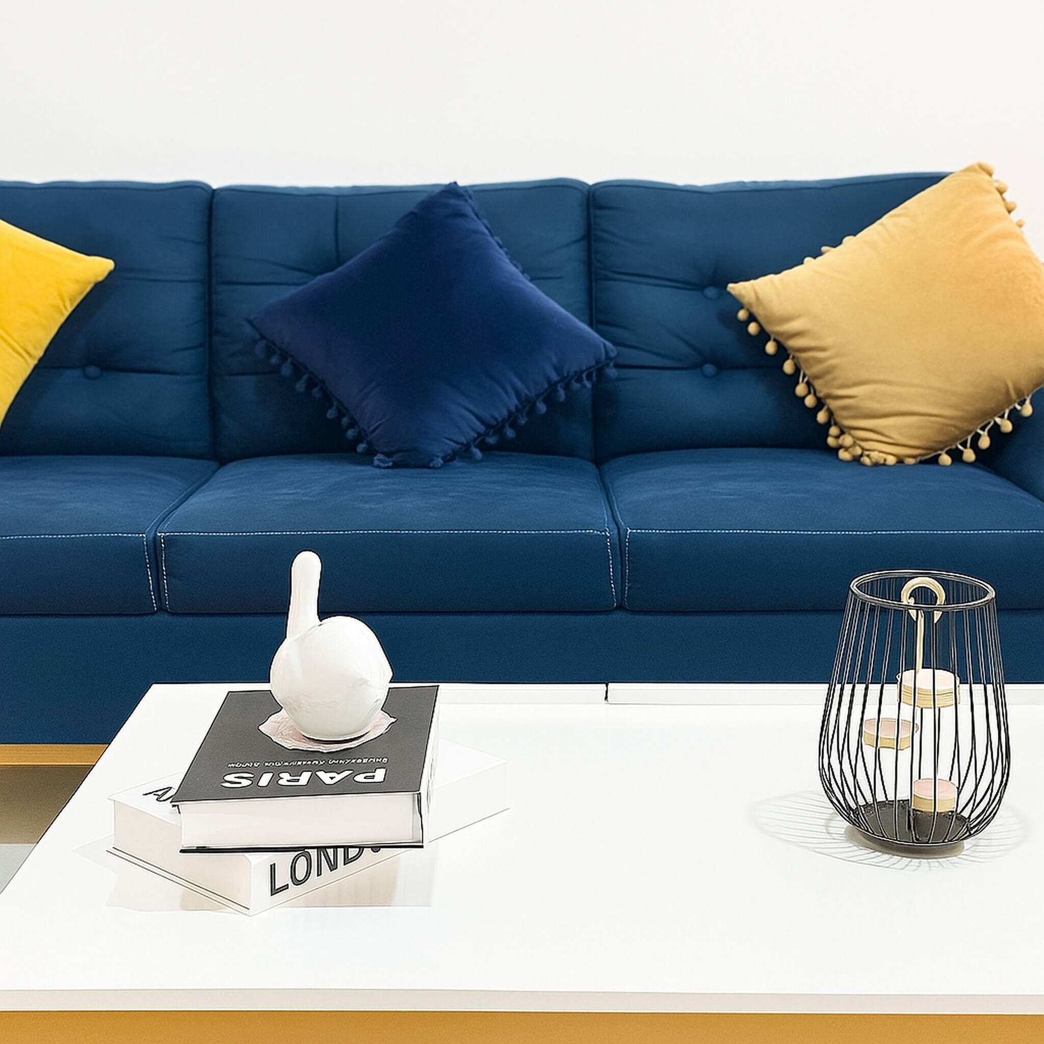 Een blauwe bank met twee kussens, een wit boek en een zwarte lamp op een witte tafel.