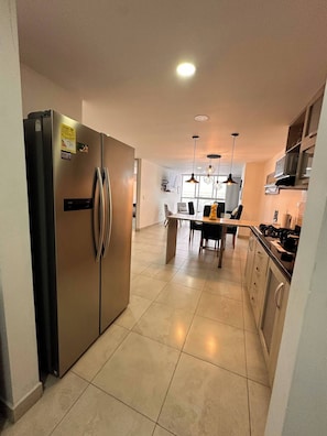 A modern kitchen with a stainless steel refrigerator, a dining table with chairs, pendant lights, and a TV mounted on the wall. - Envigado Comfortable Modern · Wifi 300 MB · Metro (Envigado)