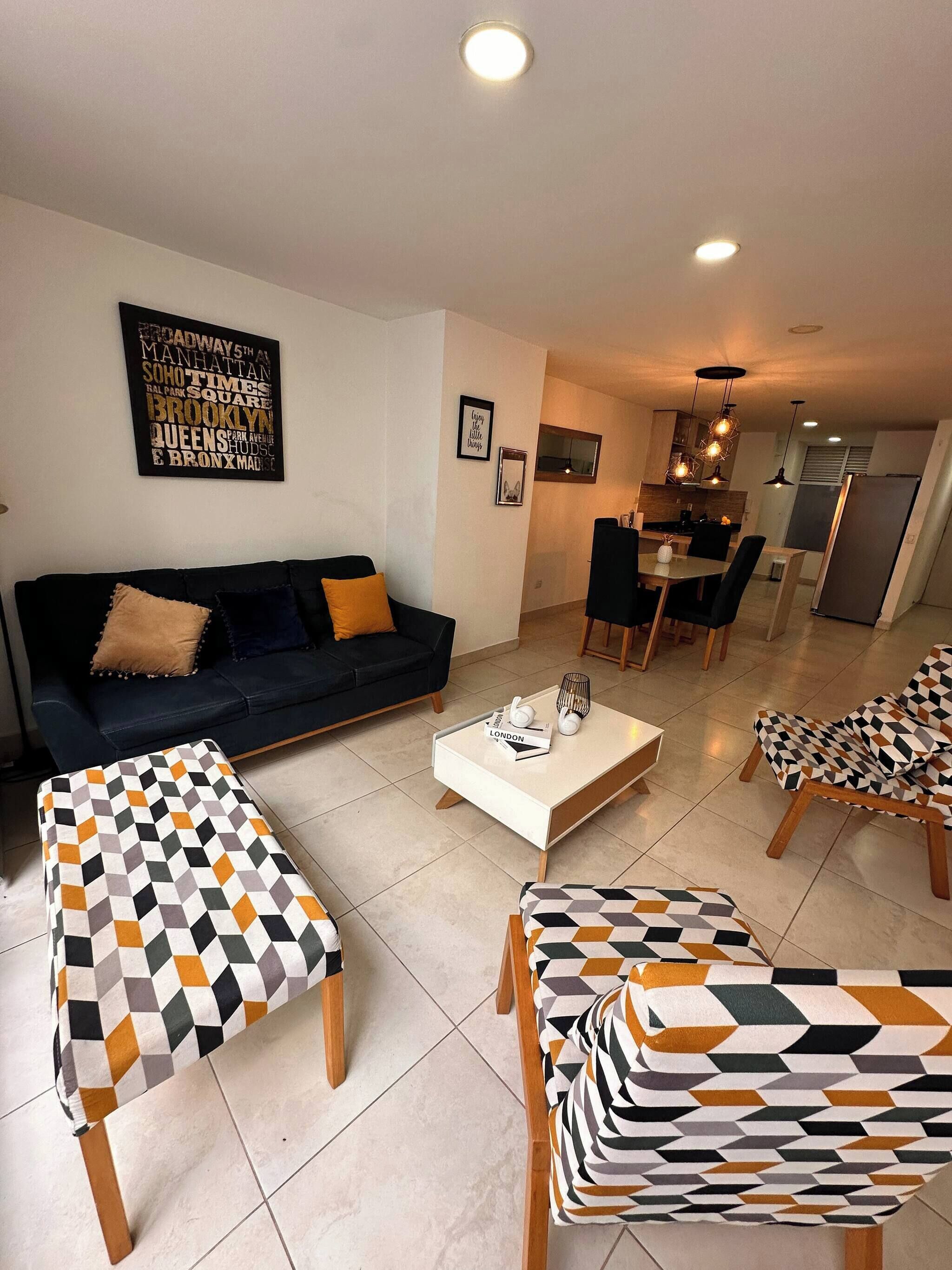 A living room with a dark sofa, patterned ottomans, a coffee table, and a dining area with chairs and a kitchen in the background.