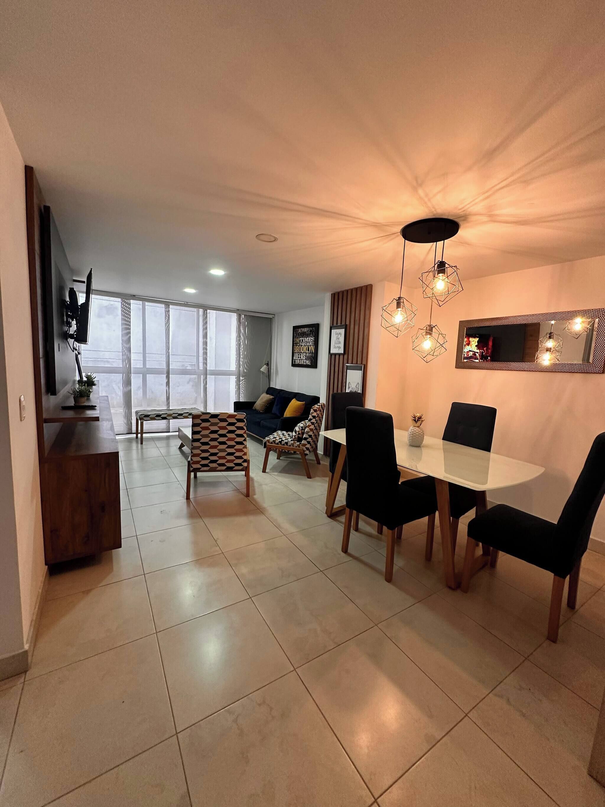 A modern living room with a sofa, a dining table with chairs, a TV mounted on the wall, and a large window.