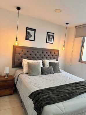 A bedroom with a tufted headboard, a bed with multiple pillows, a bedside table with a lamp, and two wall-mounted light fixtures. - Envigado Comfortable Modern · Wifi 300 MB · Metro (Envigado)