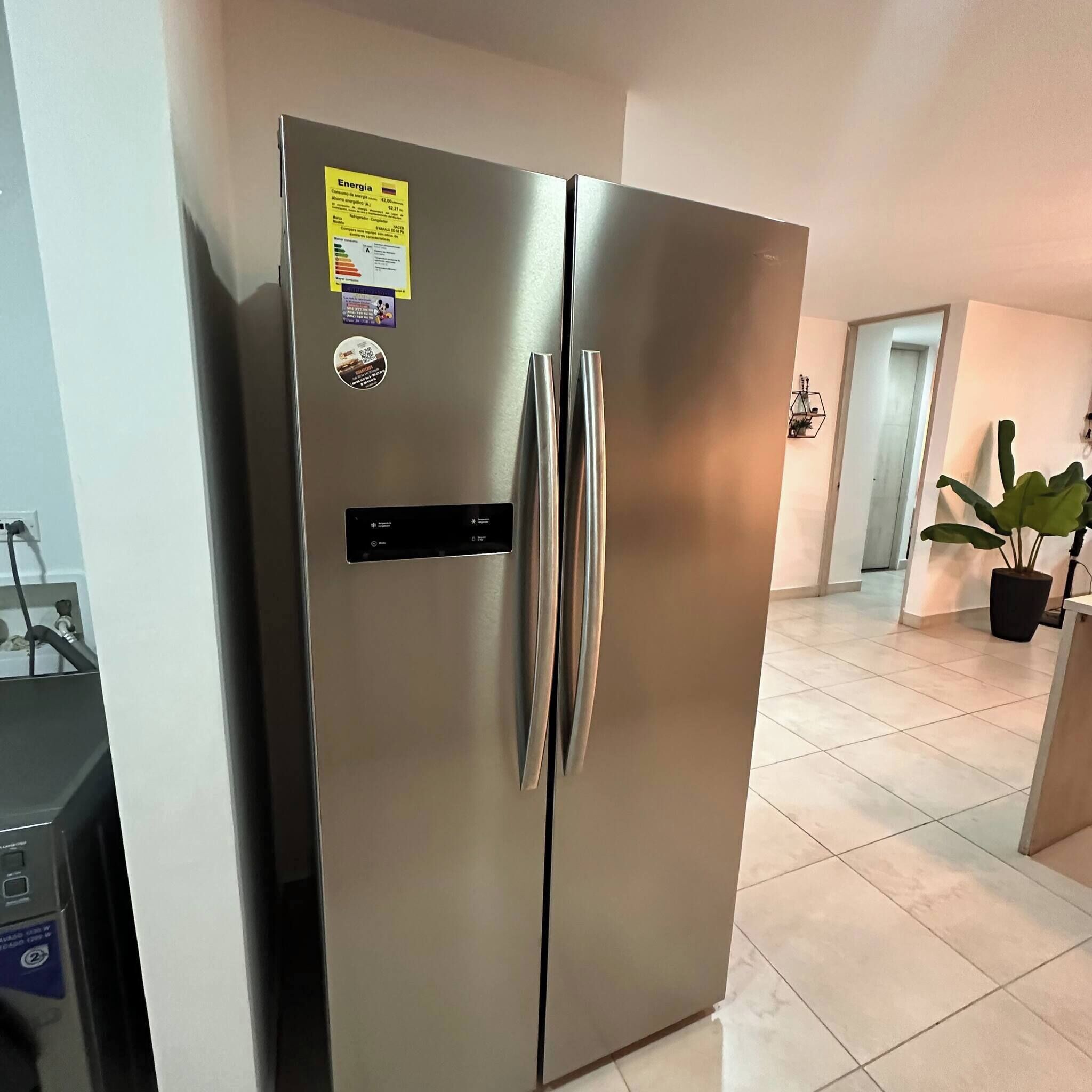 A stainless steel refrigerator with a digital control panel, located in a kitchen with tiled flooring and a potted plant.